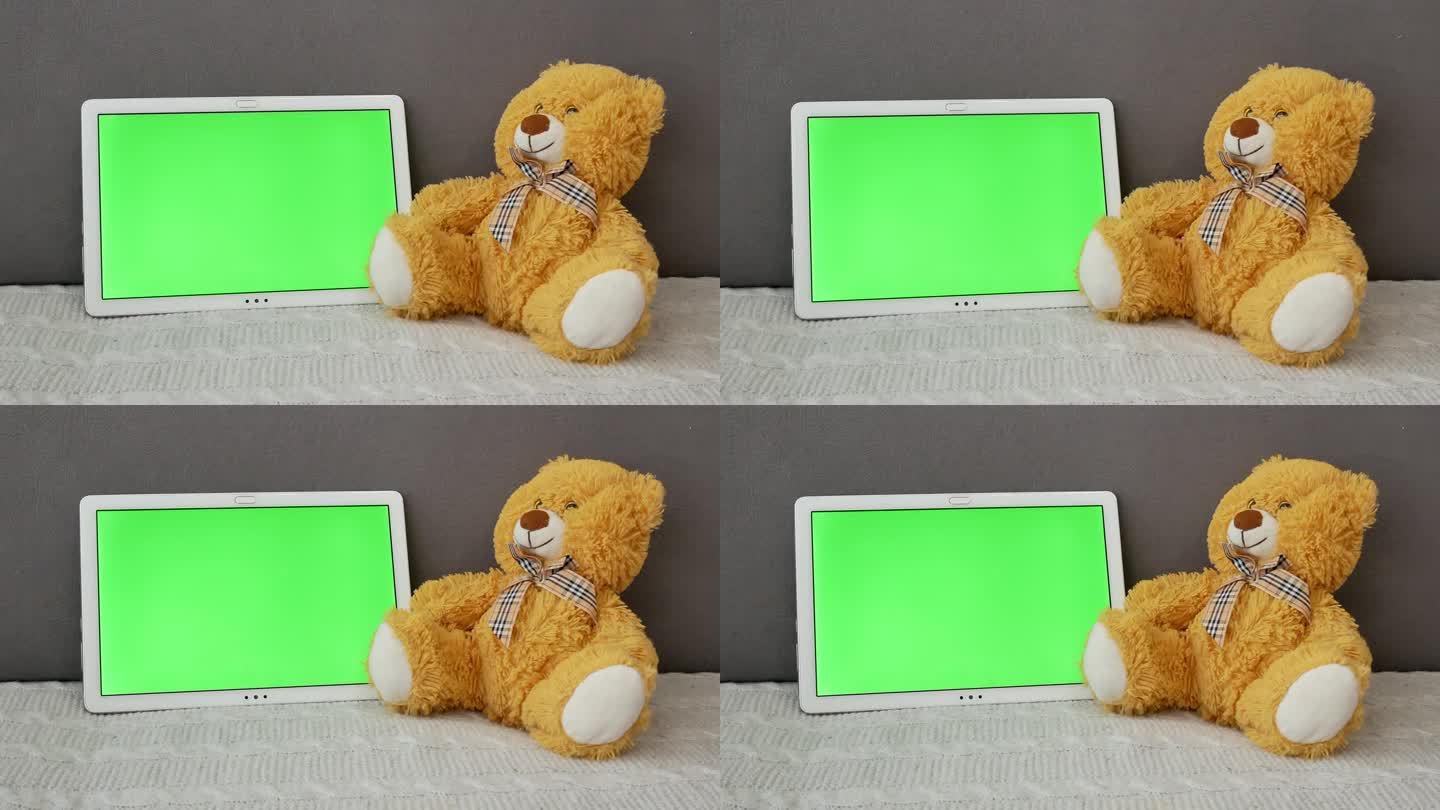 一只棕色毛绒熊坐在灰色的沙发上，旁边是一个绿色屏幕的平板电脑。科技和互联网的概念，玩具取代了电子设备