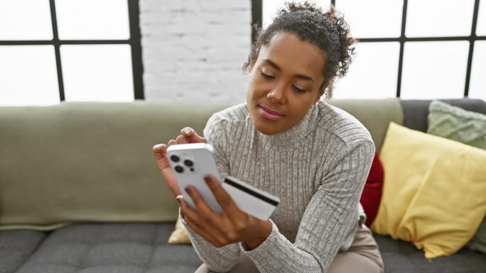 一个微笑的女人在室内用智能手机，拿着信用卡，躺在有彩色靠垫的灰色沙发上。