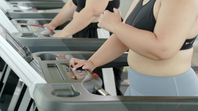 两个胖乎乎的亚洲女人穿着运动服走进跑步机。两个胖乎乎的女人都有强烈的减肥意愿、健身伙伴、生活方式和运