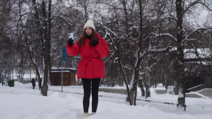 快乐漂亮的女性在下雪的冬季公园行走的全长肖像，视频用智能手机与朋友通话，讲述生活中的故事。与朋友交流