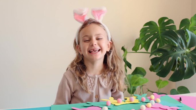 快乐的蹒跚学步的孩子笑着露出牙齿兴奋有趣惊讶的孩子女孩准备复活节聚会做纸花环彩色复活节兔子DIY。孩