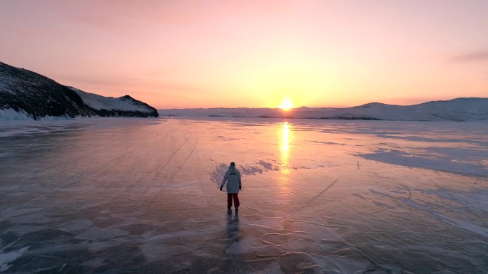 鸟瞰图上的年轻女子走在贝加尔湖的蓝色裂缝冰在美丽的橙色日出。太阳在冰面上的反射。贝加尔湖冰冻的冬季景