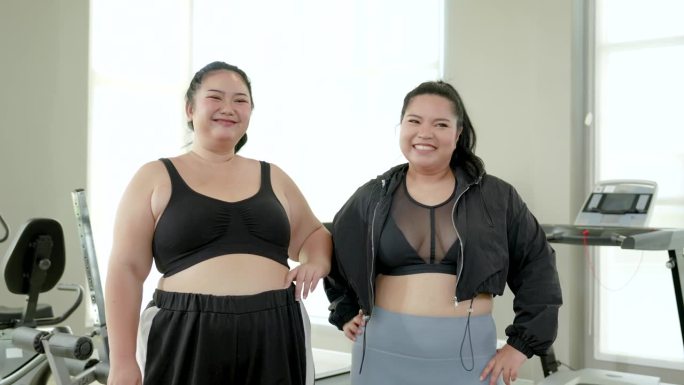 两个胖胖的亚洲女人穿着运动服，在健身房开心地微笑着。两个胖胖的女人有强烈的减肥意愿，健身伙伴，生活方