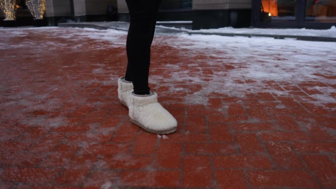 特写镜头中，一个无法辨认的女人穿着冬鞋走在滑溜溜的城市人行道上。女性沿着冰面行走的特写镜头。