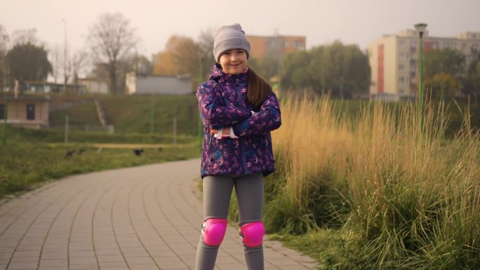在城市公园的户外，小女孩穿着护膝和护肘站在旱冰鞋上。喜欢运动的孩子