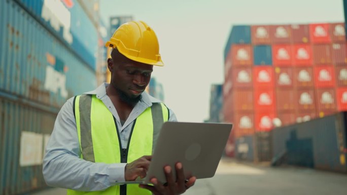 非裔美国黑人工业工程师，头戴黄色安全帽，身穿安全背心，站在集装箱堆场仓库用笔记本电脑检查集装箱。海运