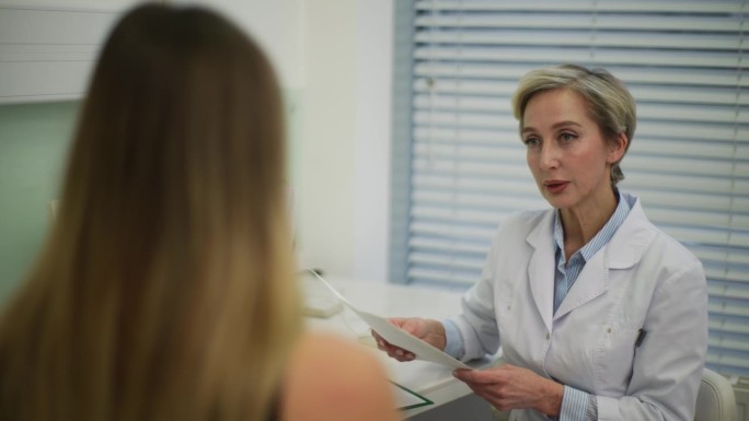 身穿白色制服、细心的中年女医生在诊所进行个人咨询时，正在给不认识的女病人开纸质处方。