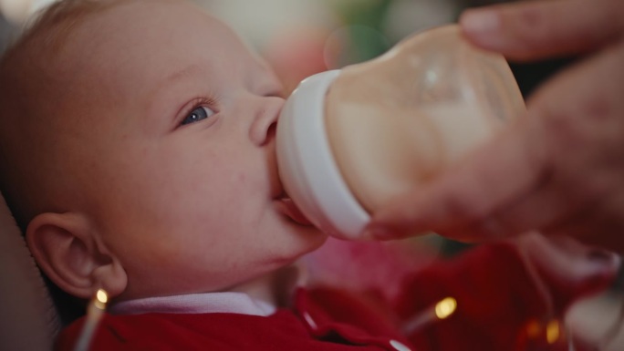 手持拍摄的顽皮的小男孩，穿着圣诞老人的服装，在家里喝着妈妈喂的奶瓶里的牛奶