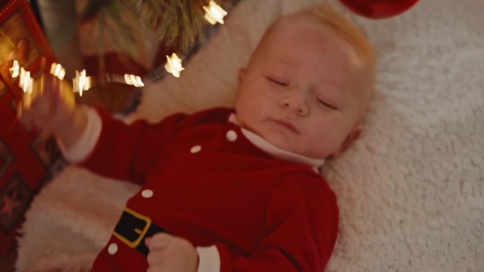 可爱的小男孩穿着圣诞老人的服装，躺在家里的地毯上玩着点亮的圣诞灯的高角度手持镜头