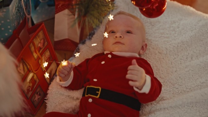 高角度手持拍摄的男婴穿着圣诞老人服装，躺在家里的地毯上玩圣诞灯
