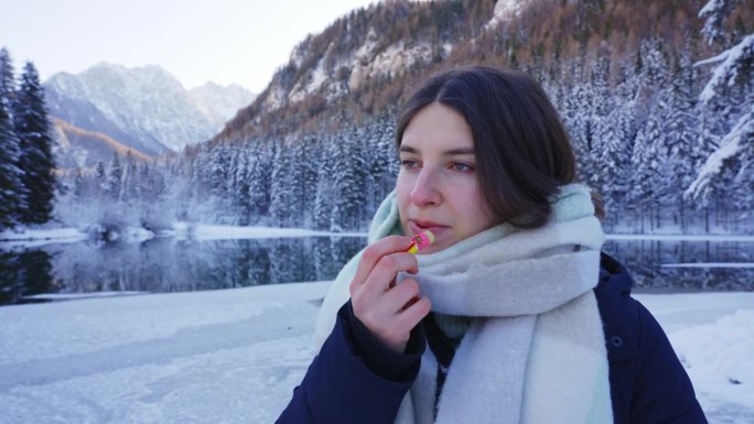 Zgornje Jezersko的雪林和山脉中，在Flozen湖附近，微笑的年轻女子涂唇膏的照片