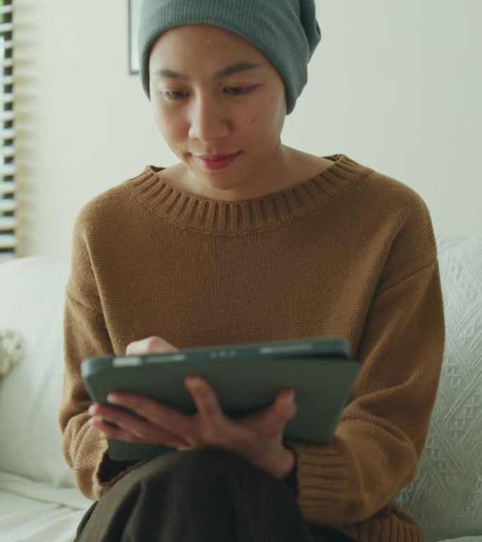 一位年轻的亚洲癌症患者戴着头巾，坐在舒适的沙发上，在光线充足的客厅里使用带触控笔的数字平板电脑。与癌
