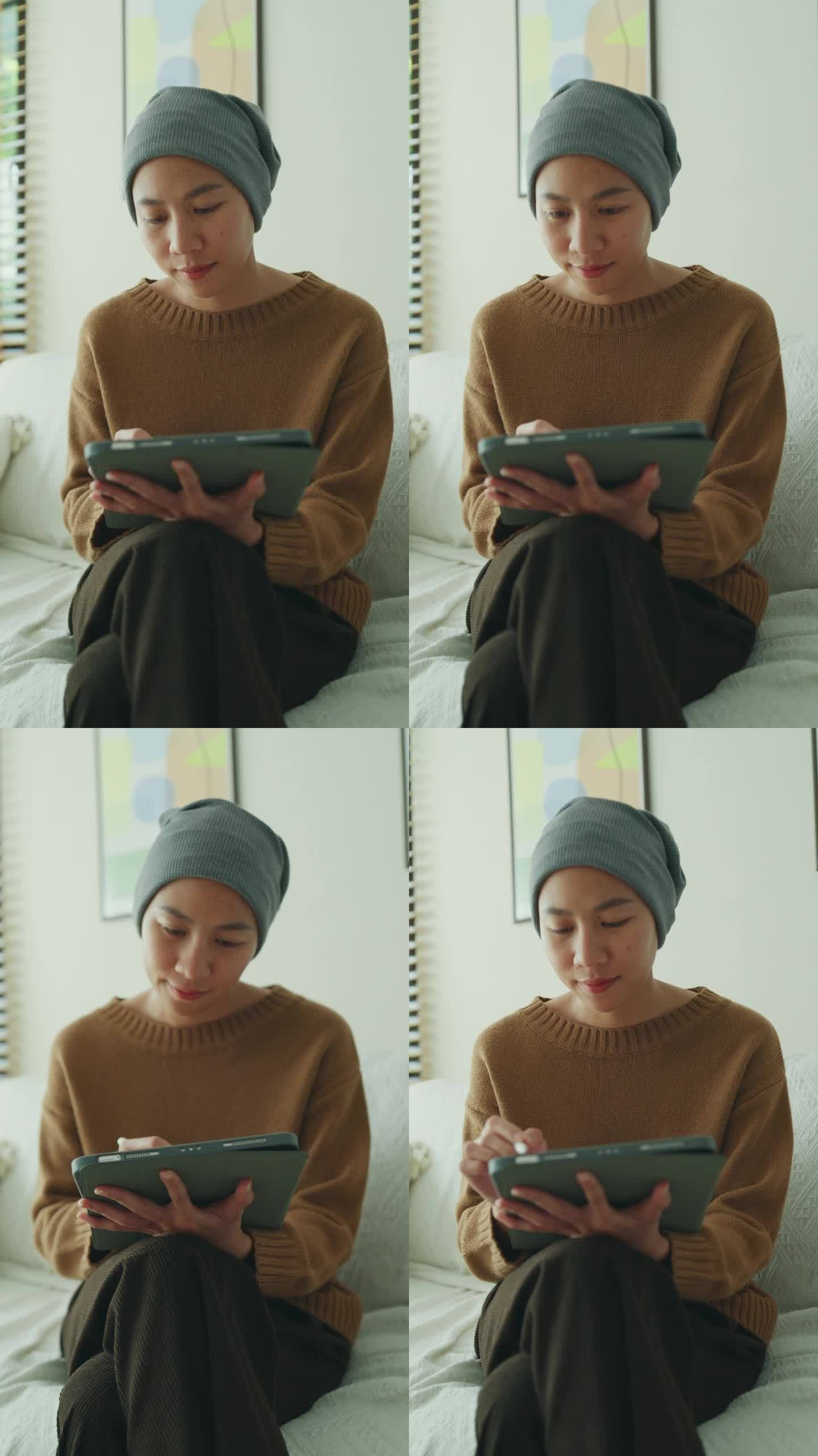 一位年轻的亚洲癌症患者戴着头巾，坐在舒适的沙发上，在光线充足的客厅里使用带触控笔的数字平板电脑。与癌