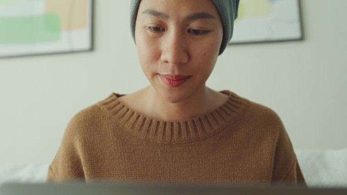 一个年轻的亚洲癌症患者，戴着头巾，舒服地坐在沙发上用笔记本电脑工作，房间里光线充足，植物装饰。与癌症