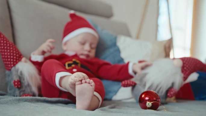 穿着圣诞老人服装的小男孩在沙发上玩小玩意和玩具的手持镜头，重点是赤脚在家