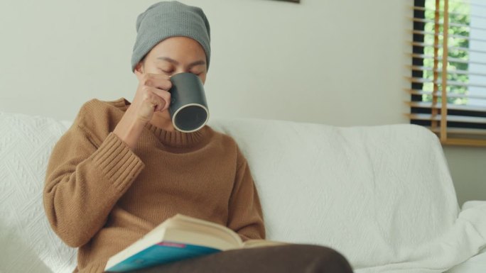 一名年轻的亚洲癌症患者戴着头巾，在阳光明媚、植物丛生的客厅里，坐在舒适的沙发上喝着咖啡，读着书。与癌