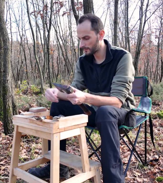木匠检查了斧头的锋利程度，开始磨它。他坐在森林里的露营椅上。