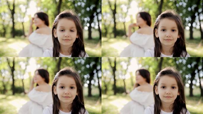 阳光下孩子的笑脸。一个女孩的肖像在前景和她的母亲在模糊的背景在公园。母女二人
