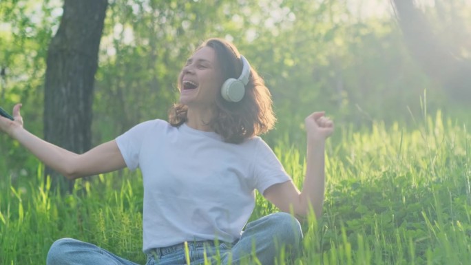 年轻美丽的兴奋女子戴着耳机，用智能手机听音乐，坐在阳光下的绿色草坪上唱歌跳舞。快乐的女性情绪。