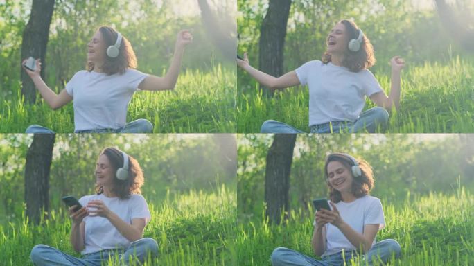 年轻美丽的兴奋女子戴着耳机，用智能手机听音乐，坐在阳光下的绿色草坪上唱歌跳舞。快乐的女性情绪。
