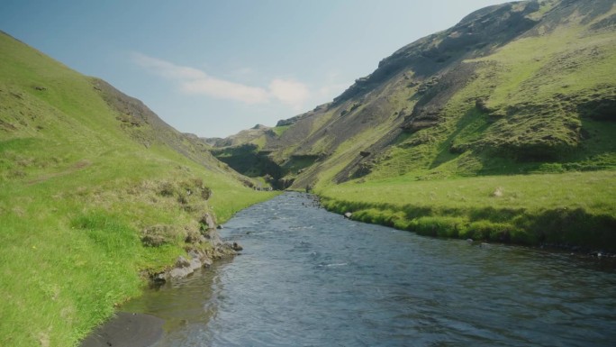 冰岛的一条小河和长满青苔的小山，前景是阳光明媚的绿色岩石和蓝色的水