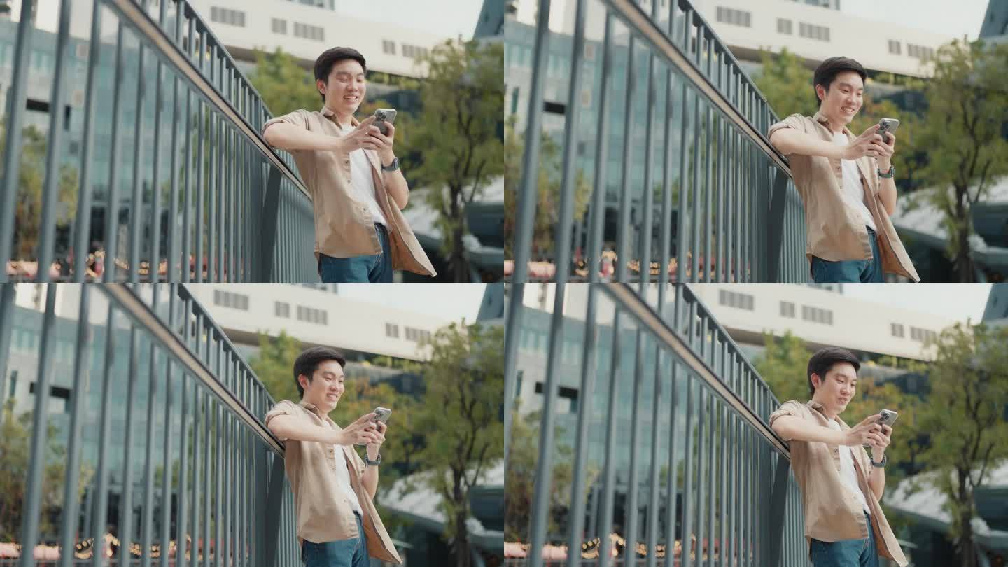 帅气的亚洲学生使用智能手机。一个年轻人站在户外，拿着手机开心地笑着