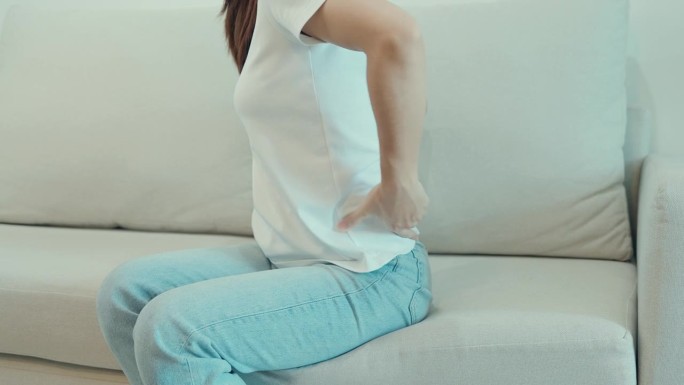 坐在家里沙发上时背部疼痛的妇女。成年女性因梨状肌综合征、腰痛和脊柱压迫引起的肌肉疼痛。办公室综合症与
