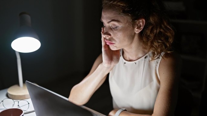 一个在办公室工作到很晚的压力很大的女人，坐在台灯下的笔记本电脑前感到筋疲力尽。