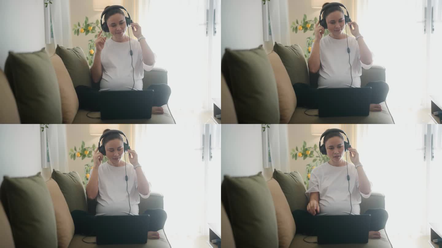 美丽快乐的孕妇坐在柔软的沙发上。在现代公寓里，用耳机和电脑工作