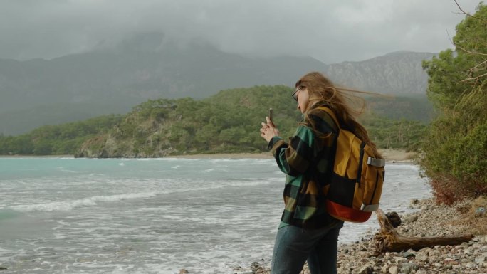 一个度假的女人，一个旅行者和博主，她的头发被海风吹乱，为社交媒体或作为纪念品拍摄了绿松石般的水和山的