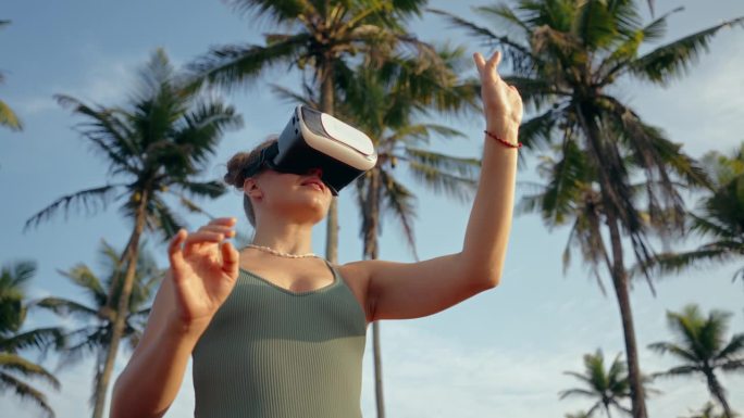 女子在日落的棕榈树中体验VR耳机，移动双手，专注于模拟。互动虚拟之旅，热带环境增强身临其境的冒险在科