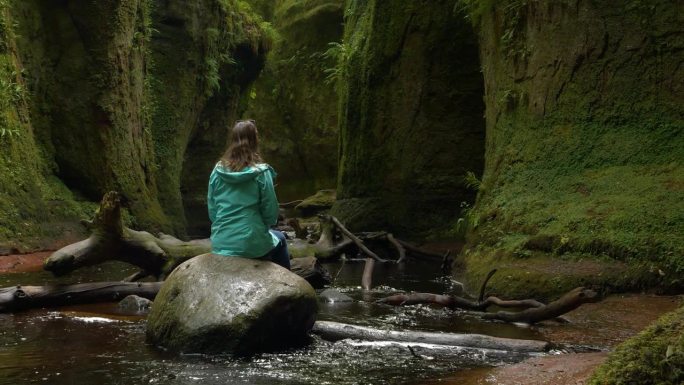年轻女子坐在翠绿的峡谷底部的一块大石头上