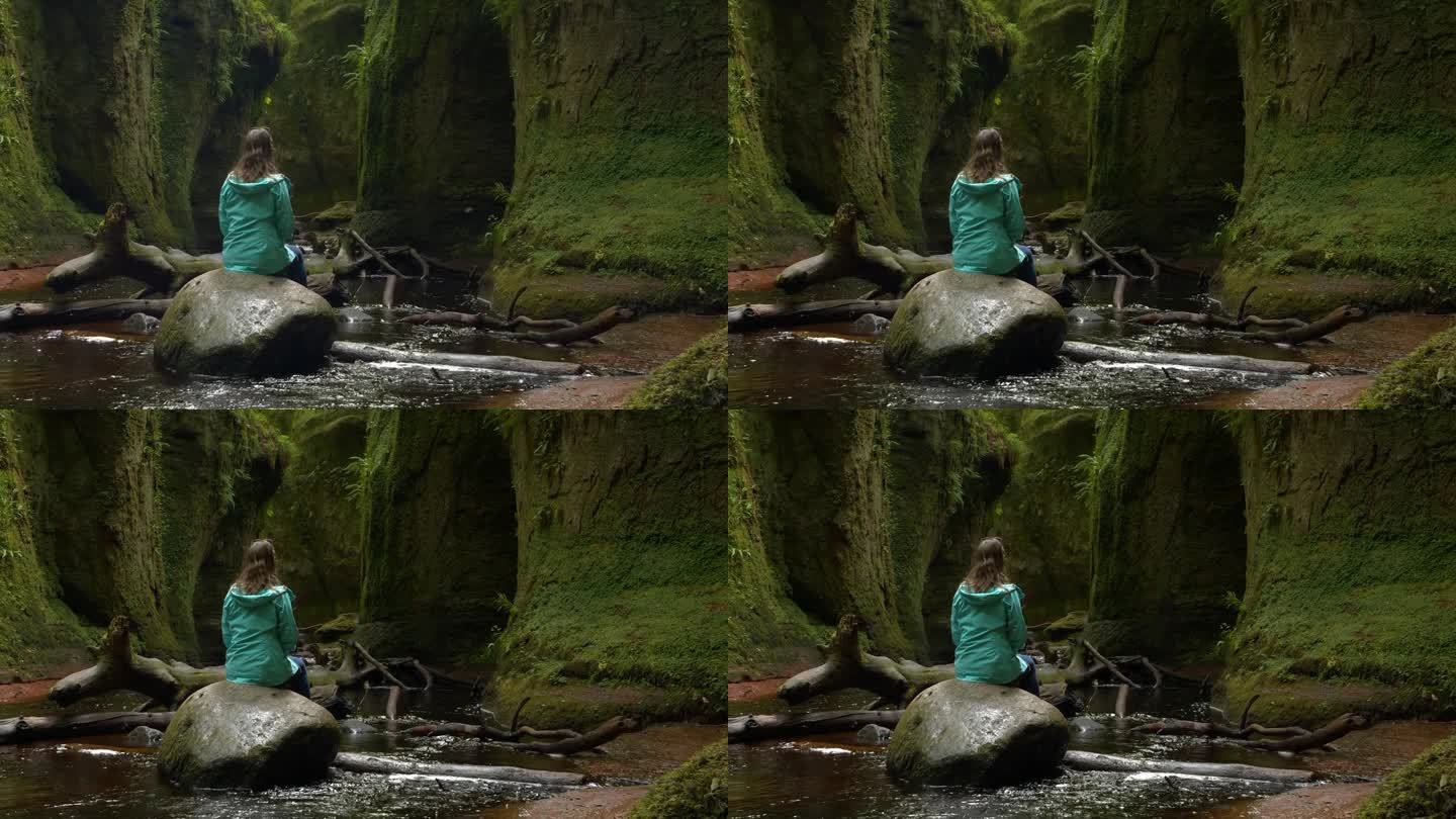 年轻女子坐在翠绿的峡谷底部的一块大石头上