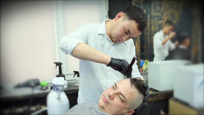 在理发店里，一位戴着黑色手套的理发师用直刀给顾客刮胡子。中景镜头。