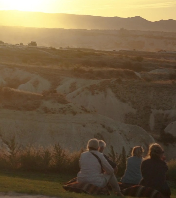 垂直视频。人们坐在山间草地上的靠垫上，看着山后温暖的夕阳。