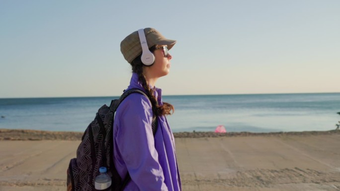 徒步旅行。一个戴着帽子、扎着小辫、背着背包的年轻女子，在沙滩上沿着海边散步，戴着耳机听音乐。侧视图，