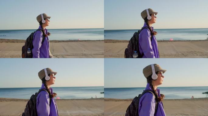 徒步旅行。一个戴着帽子、扎着小辫、背着背包的年轻女子，在沙滩上沿着海边散步，戴着耳机听音乐。侧视图，