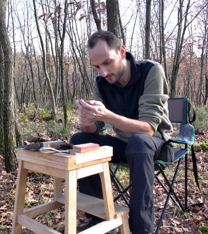 一个人站在木雕椅上，坐在满是秋叶的森林里的露营椅上雕刻木雕。