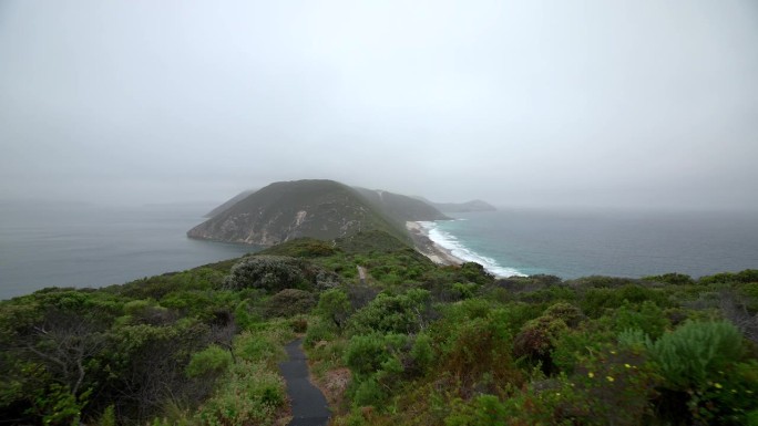 在一个雾蒙蒙的日子里，镜头沿着一条风景优美的小路向前推进，沿着一条狭窄的岬角延伸到海洋。
