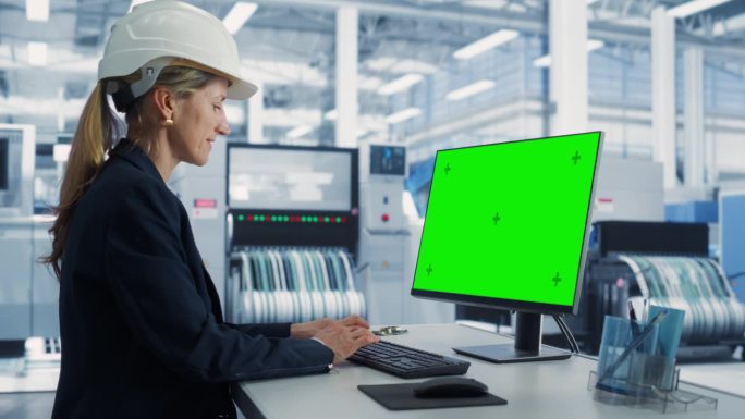 一名白人女性软件开发人员在一家工厂的台式电脑上工作，电脑上显示的是绿屏铬色。技术研发办公室监控生产