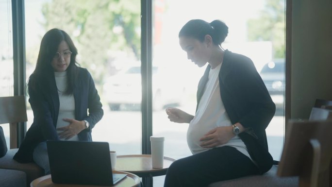 两名孕妇在咖啡馆工作的特写镜头，她用笔记本电脑抚摸着自己的肚子。