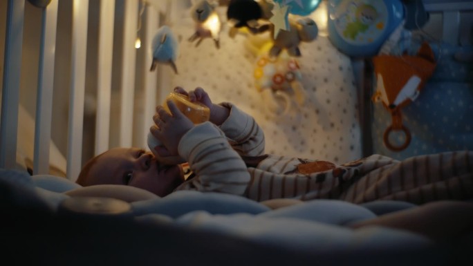 在家里的暗房里，可爱的小男孩躺在婴儿床上喝着奶瓶里的牛奶。婴儿床里的男婴