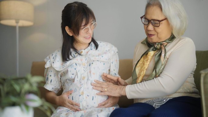 亚洲老年妇女抚摸怀孕女儿的肚子，在客厅与女儿交谈。