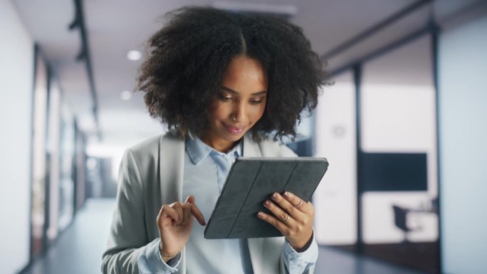 女性社交媒体协调员微笑着在平板电脑上查看新数据。一位成功的非洲女商人走在宽敞的公司办公楼里的肖像
