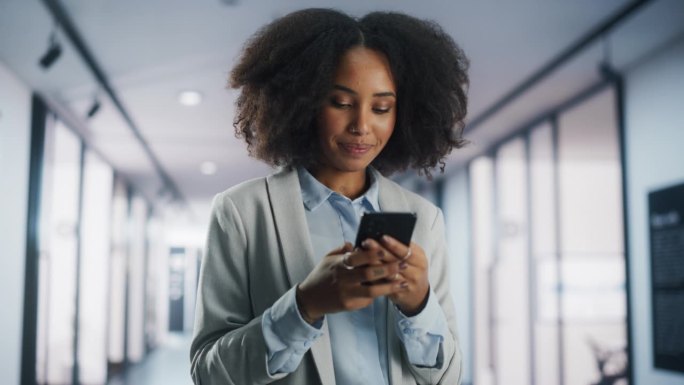 一位快乐的非裔美国女性站在公司办公室里用智能手机发短信，微笑着。年轻黑人女性市场经理查看社交网络，与