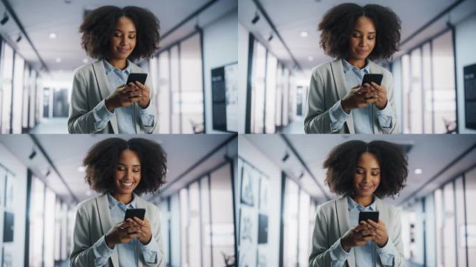 一位快乐的非裔美国女性站在公司办公室里用智能手机发短信，微笑着。年轻黑人女性市场经理查看社交网络，与