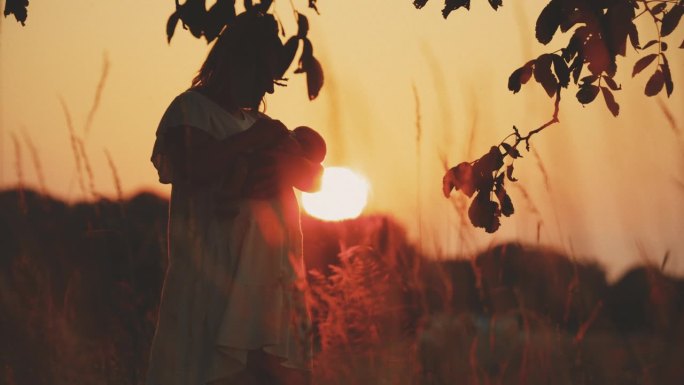 黄昏时分，站在橙色天空的田野上，快乐的母亲抱着婴儿男孩的SLO手持镜头