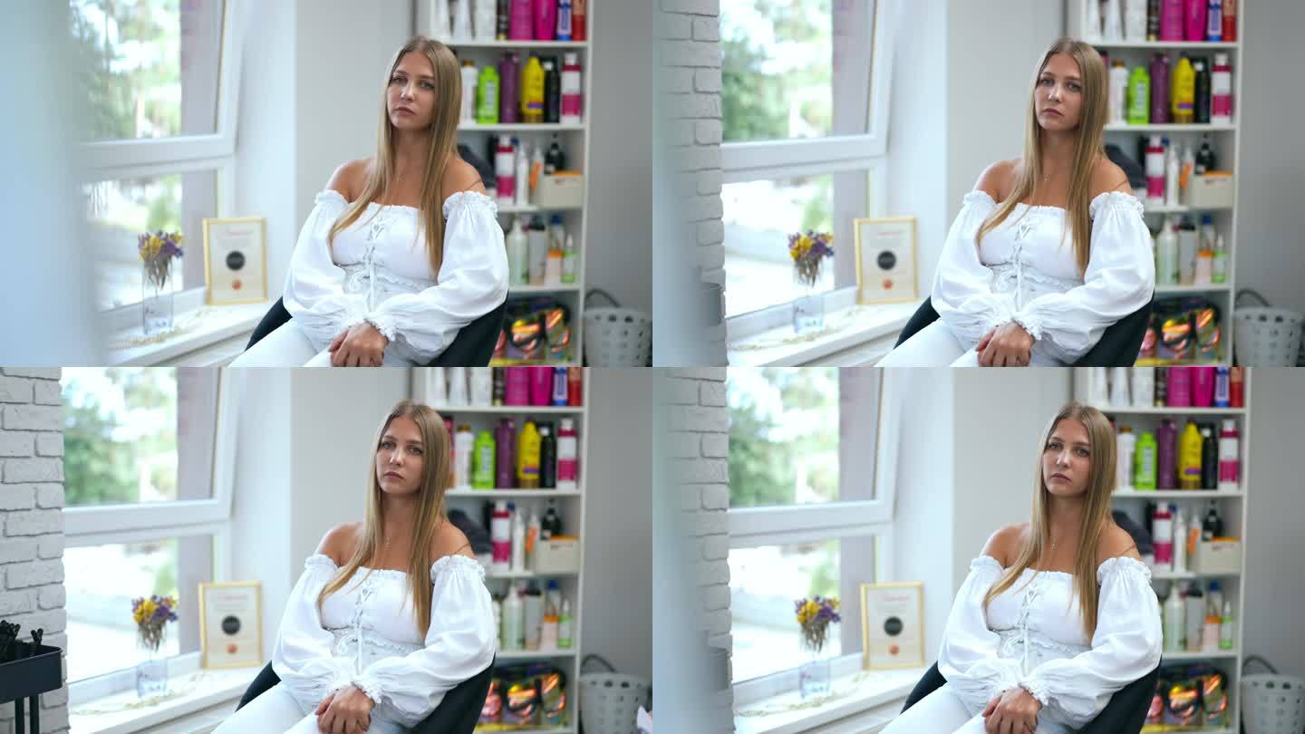 在一家美容院里，一个漂亮的女孩坐在靠窗的椅子上，背景是一个护发用品摊位。