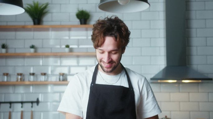 微笑的卷发视频博主男厨师在围裙录制视频烹饪课素食餐在现代厨房。一个接一个地展示蔬菜