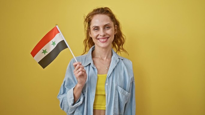 面带微笑的年轻女子手持一面叙利亚国旗，靠着一面黄色的墙，展现出积极向上和文化自豪感。